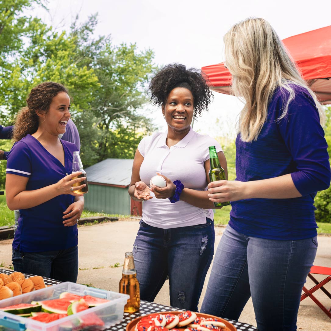 Women smiling while they network outdoors in front of a table full of food.